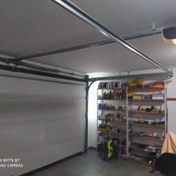 Berlin-Technik Łukasz Berliński - Otwarta brama garażowa segmentowa z widocznym mechanizmem podnoszenia i półkami z narzędziami w tle.