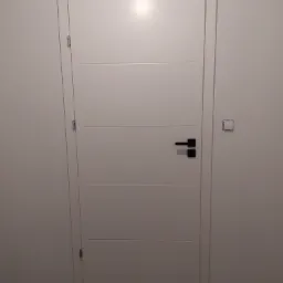 Białe drzwi wewnętrzne z poziomymi frezami i czarną klamką, zamontowane w białej ścianie, widoczny fragment drewnianej podłogi i niezamaskowany włącznik światła.