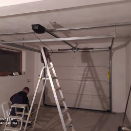 Berlin-Technik Łukasz Berliński - Proces instalacji bramy garażowej: widoczny mechanizm napędowy pod sufitem, drabina aluminiowa oraz pracownik w trakcie montażu elementów konstrukcyjnych w surowym garażu.
