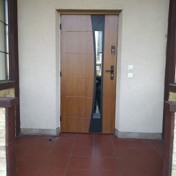 Berlin-Technik Łukasz Berliński - Zewnętrzne drzwi wejściowe z panelem bocznym w kolorze czarnym i szybą, z brązowymi poziomymi panelami na drewnopodobnej powierzchni, zamontowane w beżowej ścianie budynku z brązowymi ramami...