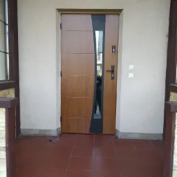 Zewnętrzne drzwi wejściowe z panelem bocznym w kolorze czarnym i szybą, z brązowymi poziomymi panelami na drewnopodobnej powierzchni, zamontowane w beżowej ścianie budynku z brązowymi ramami...