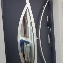 Berlin-Technik Łukasz Berliński - Grafitowe drzwi zewnętrzne z dekoracyjnym panelem szklanym w kształcie łzy, wykończone srebrnymi elementami, z długim, zakrzywionym uchwytem i domofonem, osadzone w surowym, betonowym otworze.