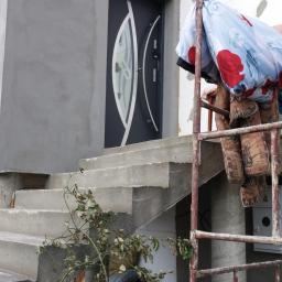 Berlin-Technik Łukasz Berliński - Szare drzwi zewnętrzne z dekoracyjnym przeszkleniem, widoczne z dołu schodów betonowych podczas prac wykończeniowych elewacji budynku, z rusztowaniem i wiszącymi materiałami budowlanymi.