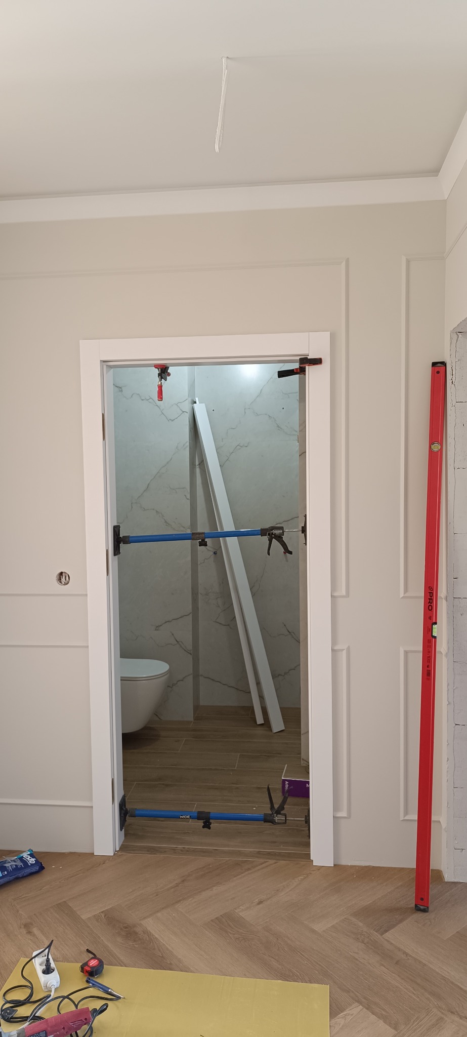 Białe drzwi wewnętrzne w trakcie montażu, widoczne narzędzia i poziomica. W tle wykończona łazienka z marmurowymi płytkami i toaletą. Parkiet ułożony w jodełkę.