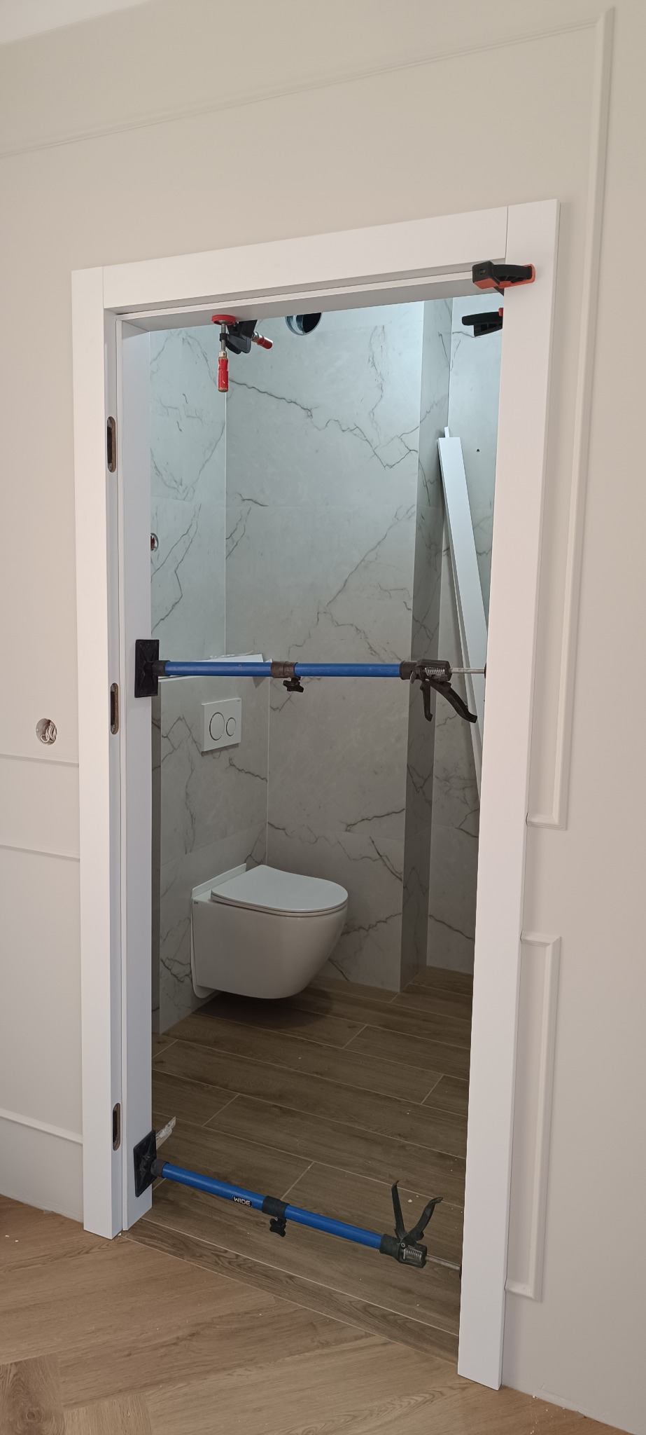 Białe ościeżnice drzwiowe w trakcie montażu, podparte niebieskimi rozpórkami. W tle widoczna toaleta z marmurowymi płytkami. Ujęcie z bliska, widać detale wykończenia.