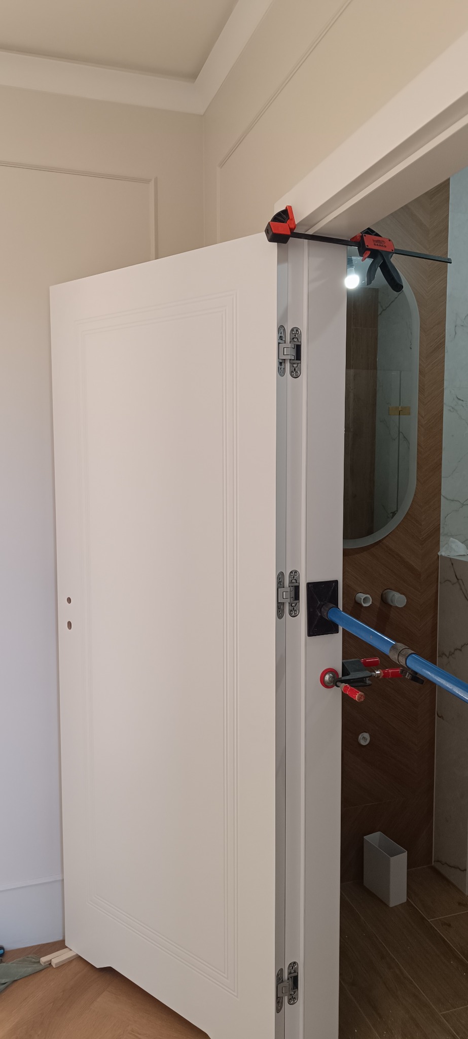 Białe drzwi wewnętrzne w trakcie montażu, widoczne zawiasy i narzędzia monterskie. W tle łazienka z owalnym lustrem i drewnianą okładziną.