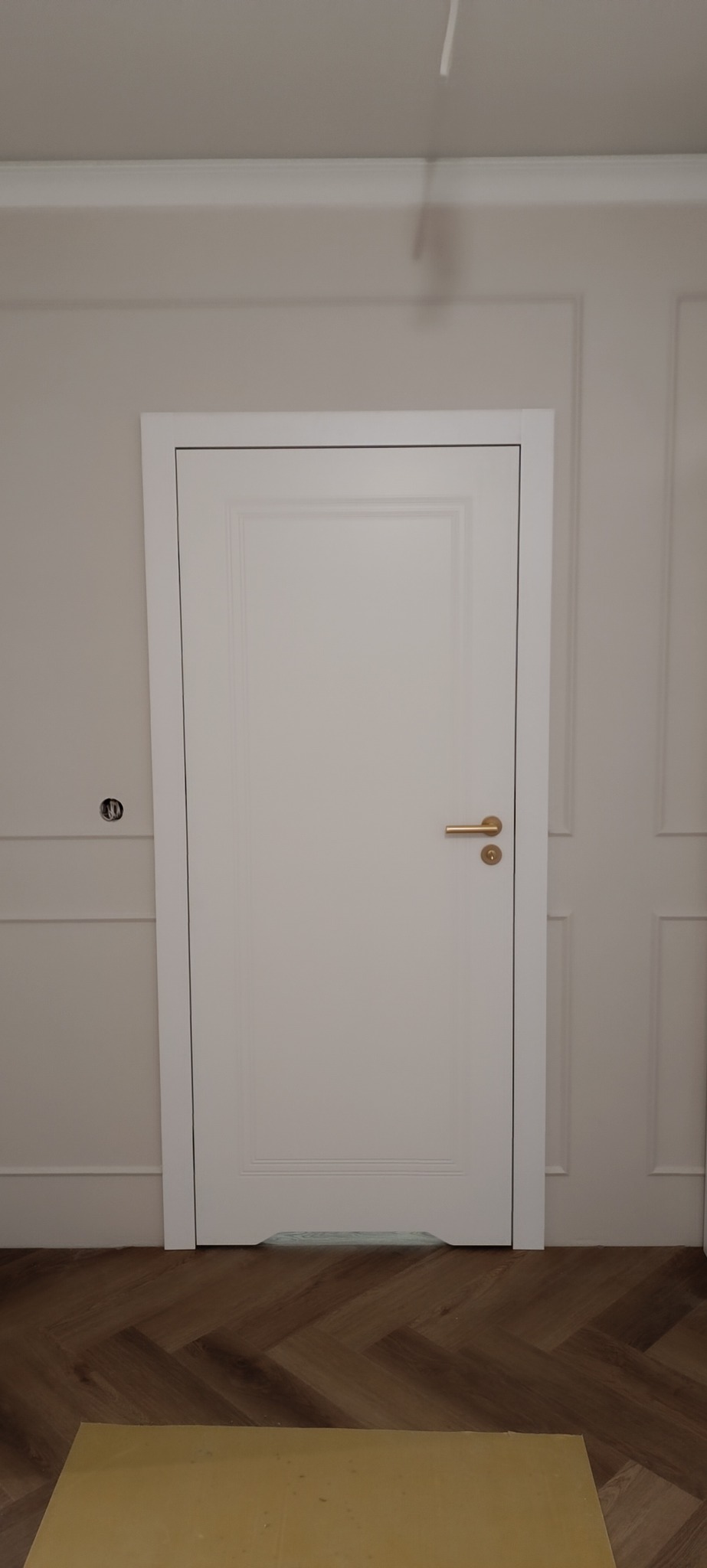 Białe, klasyczne drzwi wewnętrzne z delikatnym frezowaniem i złotą klamką, zamontowane w ścianie z dekoracyjnymi listwami. Parkiet w jodełkę na podłodze.