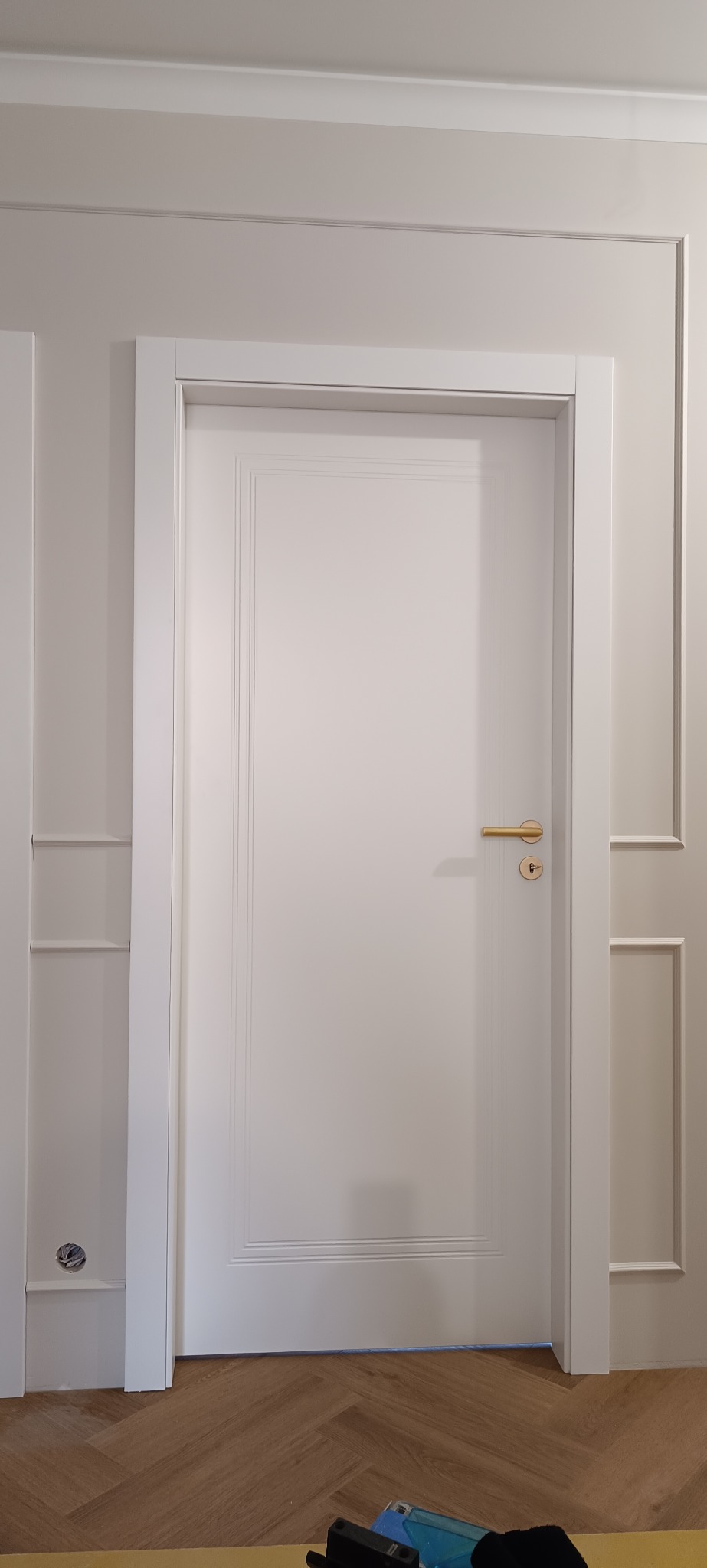 Białe drzwi wewnętrzne z złotą klamką, osadzone w białej futrynie. Ściana z dekoracyjnymi listwami. Parkiet ułożony w jodełkę. Widoczne narzędzia na podłodze.