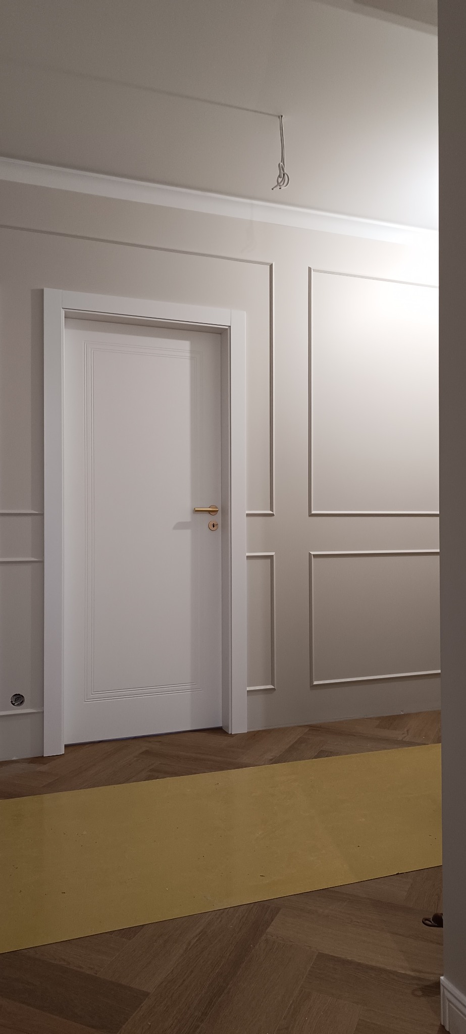 Białe drzwi wewnętrzne z złotą klamką w eleganckim wnętrzu z panelami ściennymi i parkietem w jodełkę. Widoczne przewody oświetleniowe na suficie.