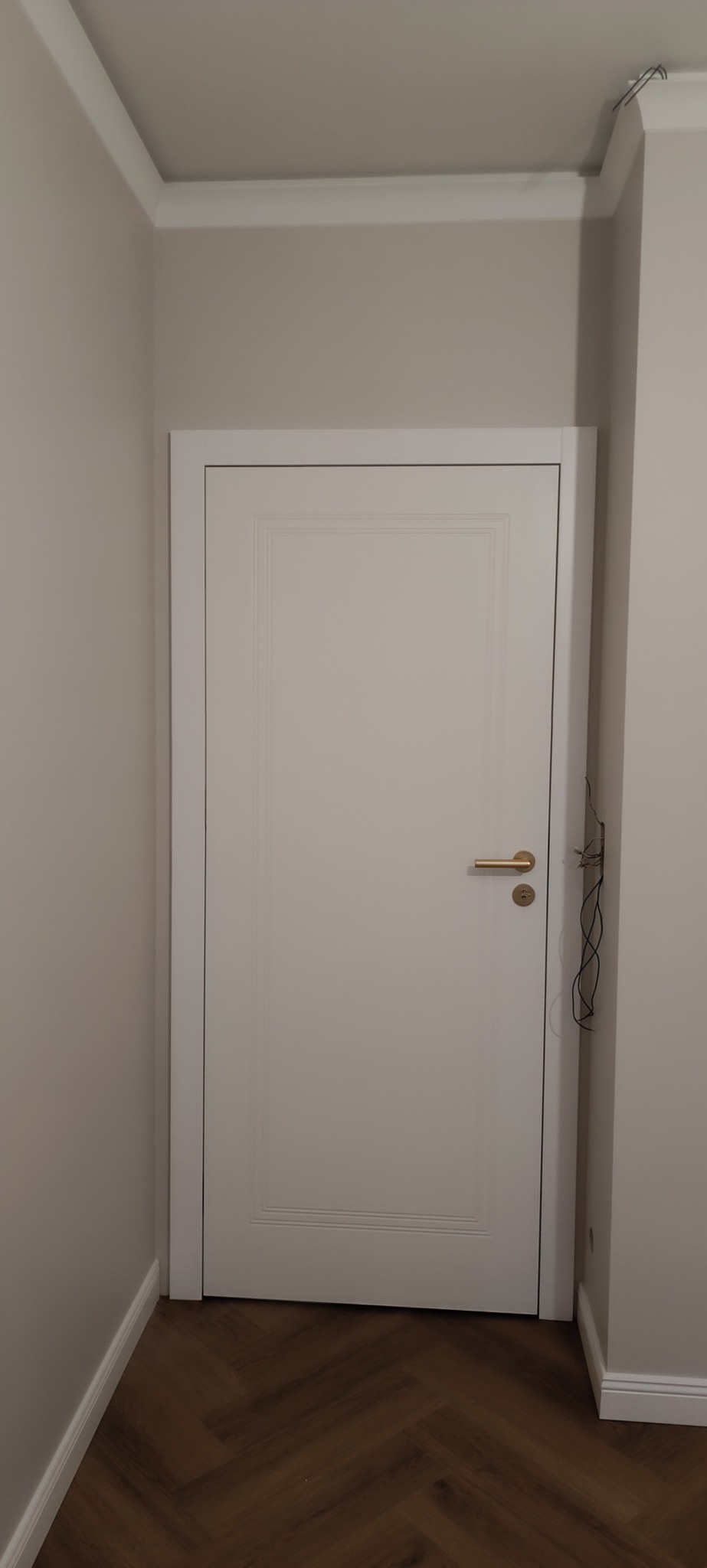 Zamknięte, białe drzwi wewnętrzne z złotą klamką, w jasnym pomieszczeniu z parkietem ułożonym w jodełkę. Widoczne listwy przypodłogowe i fragment instalacji elektrycznej.