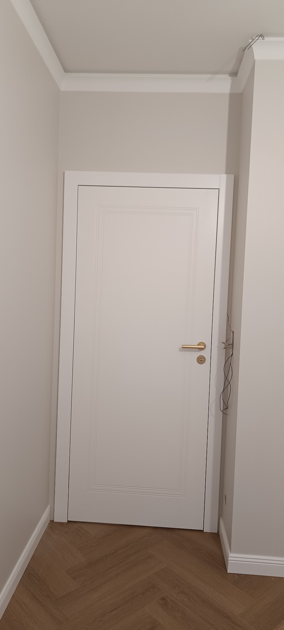 Białe drzwi wewnętrzne z delikatnym frezowaniem i złotą klamką, zamontowane w jasnym pomieszczeniu z parkietem ułożonym w jodełkę. Widoczne przewody wystające ze ściany.