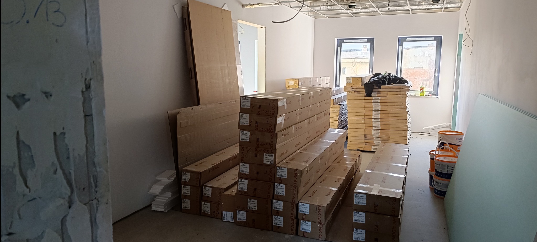 Wnętrze w trakcie remontu: stosy zapakowanych drzwi, płyty gipsowe i farby. Widoczne okna i sufit w trakcie prac wykończeniowych. Przygotowanie do montażu.