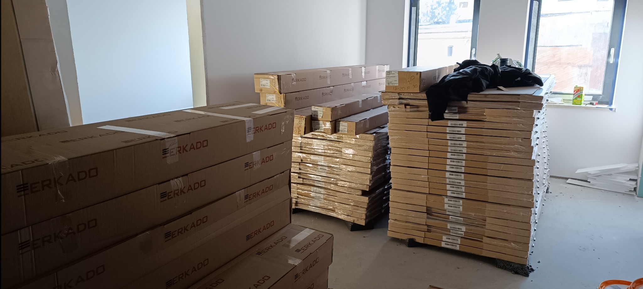 Stosy zapakowanych drzwi marki ERKADO w pomieszczeniu przed montażem. Widoczne kartony i folie ochronne, przygotowane do instalacji w nowym wnętrzu.