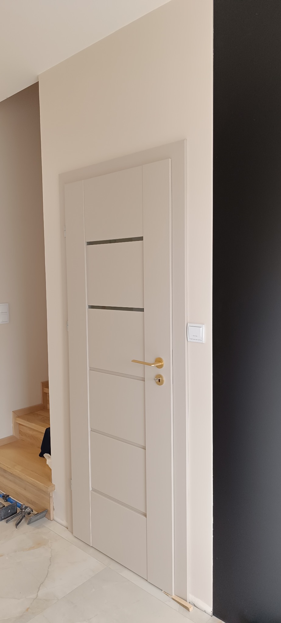 Beżowe drzwi wewnętrzne z poziomymi, ciemnymi paskami i złotą klamką, zamontowane w jasnym pomieszczeniu z widocznymi schodami i narzędziami.