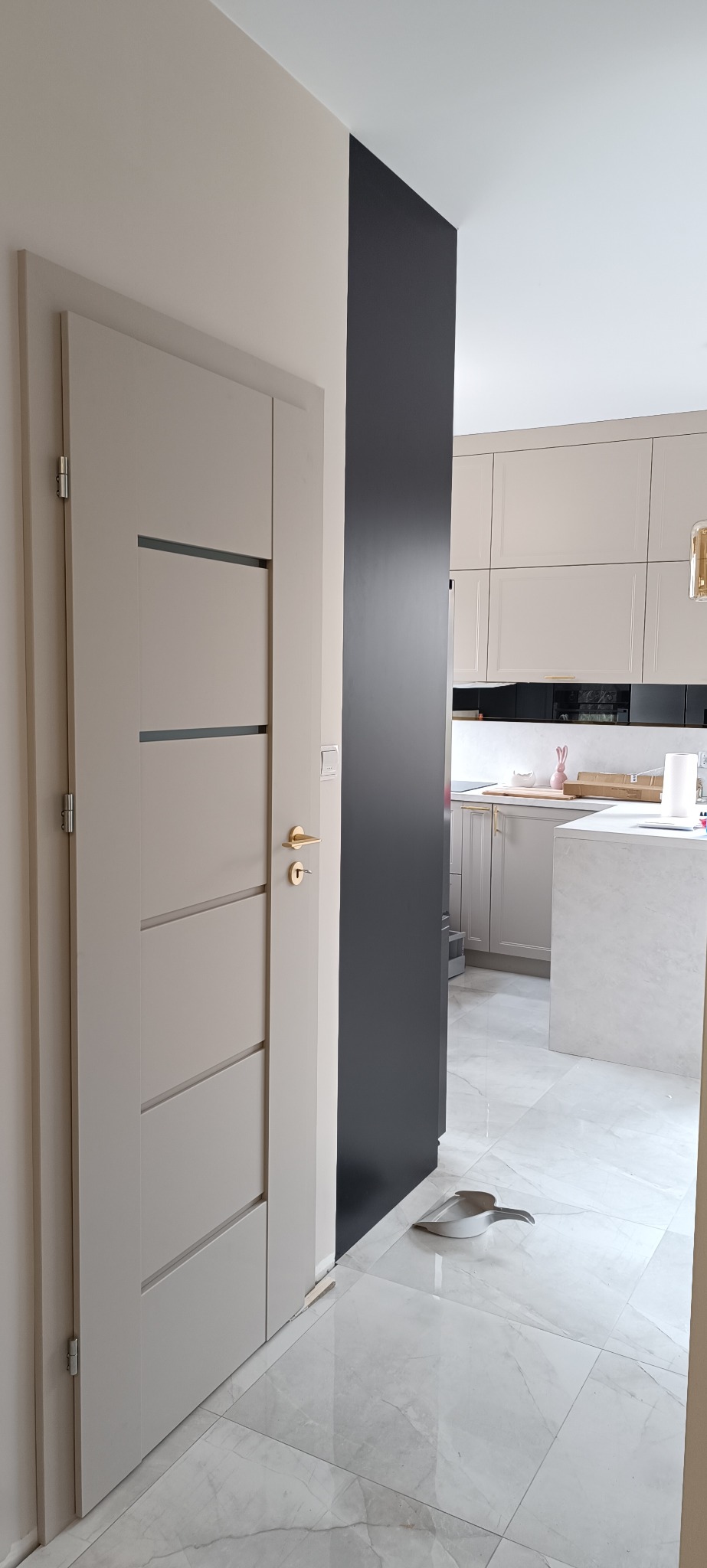 Beżowe drzwi wewnętrzne z poziomymi przeszkleniami i złotą klamką, zamontowane w mieszkaniu w Makowie Mazowieckim. Widok na kuchnię z marmurową wyspą.