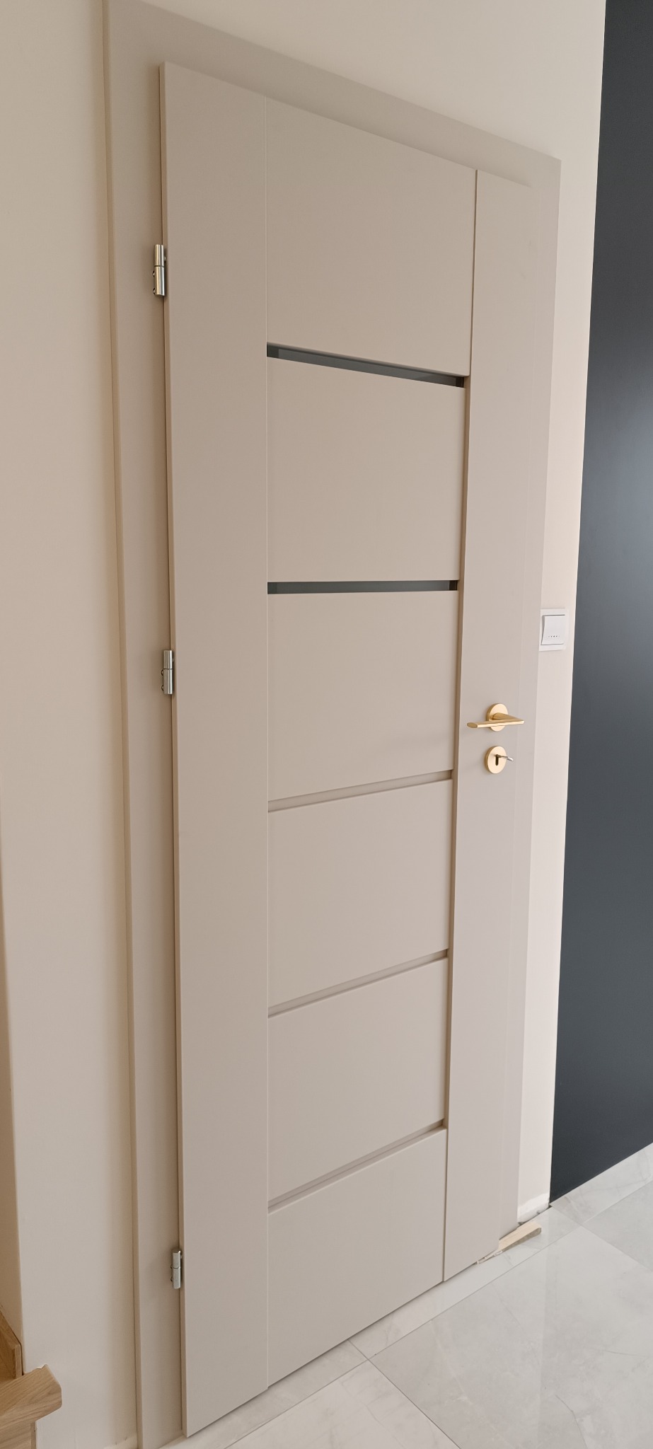 Beżowe drzwi wewnętrzne z poziomymi, ciemnymi wstawkami i złotą klamką, zamontowane w jasnym pomieszczeniu z marmurową podłogą i fragmentem ciemnej ściany.
