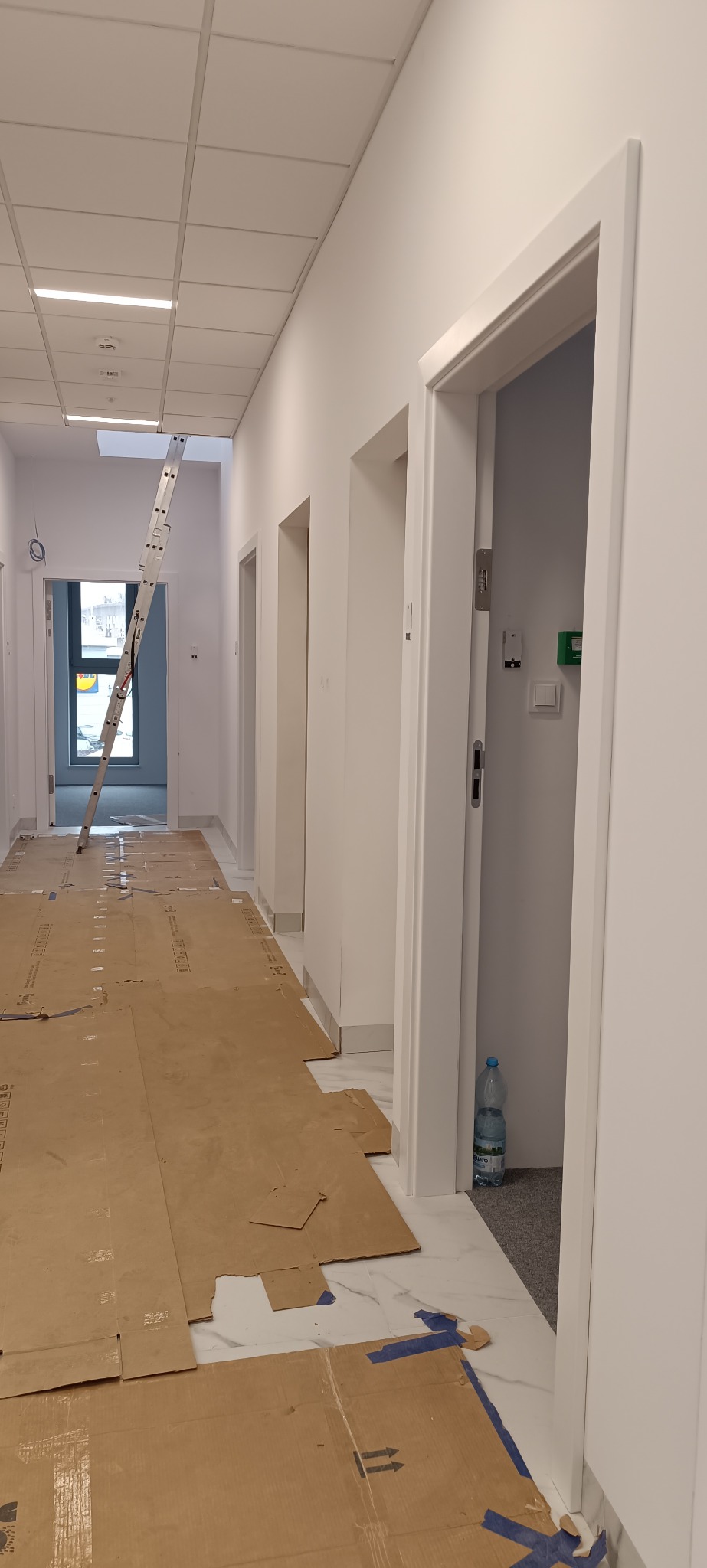 Korytarz w trakcie remontu z zabezpieczoną podłogą kartonem i widoczną drabiną opartą o sufit. Białe ściany i ościeżnice drzwiowe.