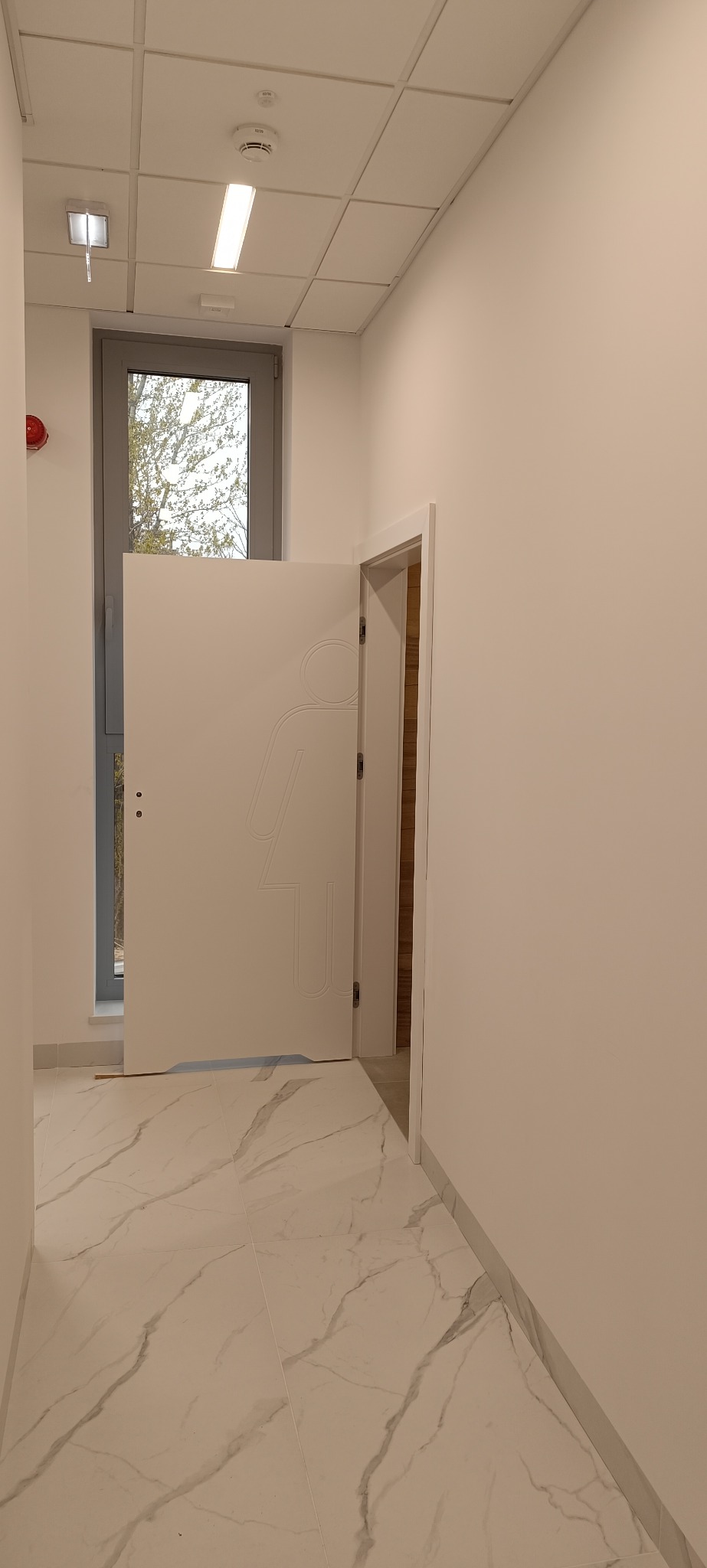 Białe drzwi z symbolem kobiety, lekko uchylone, w jasnym korytarzu z marmurową podłogą. Widoczne okno z widokiem na drzewo. Minimalistyczny design.