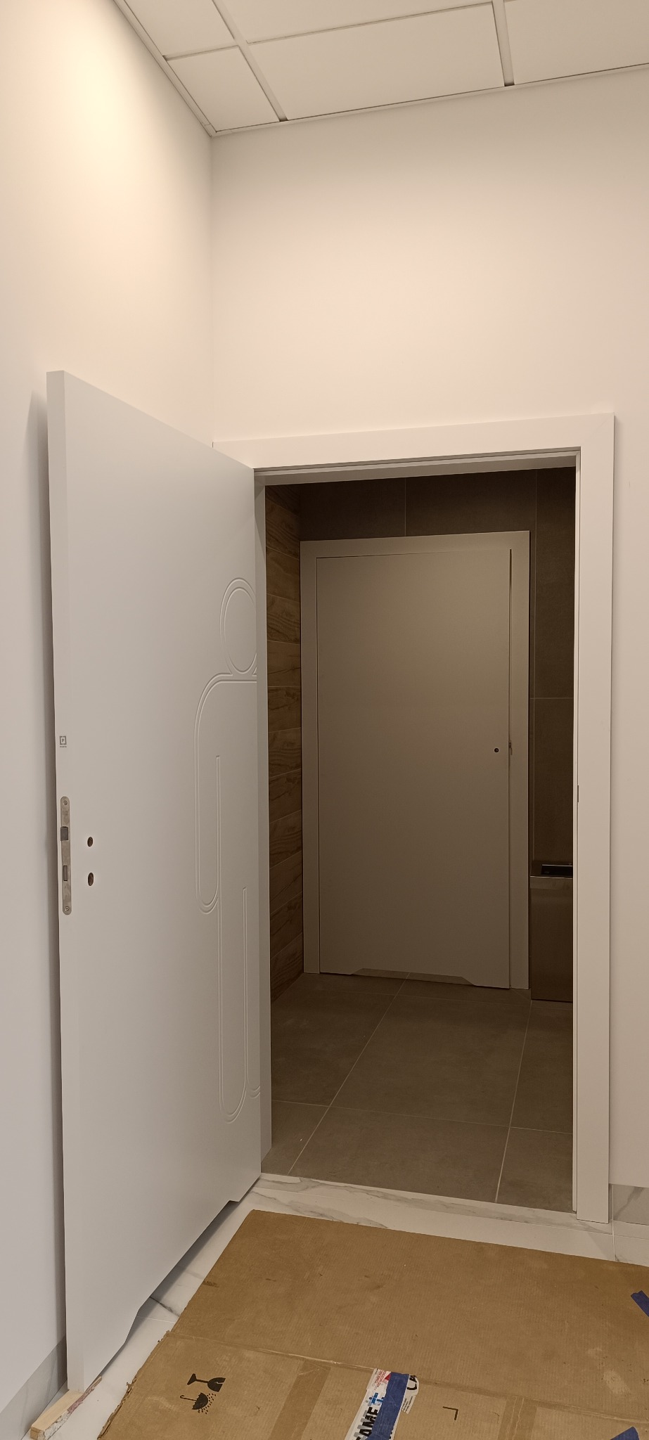 Białe, częściowo otwarte drzwi wewnętrzne z dekoracyjnym frezem, widoczne w przejściu do innego pomieszczenia z szarą podłogą i drzwiami.