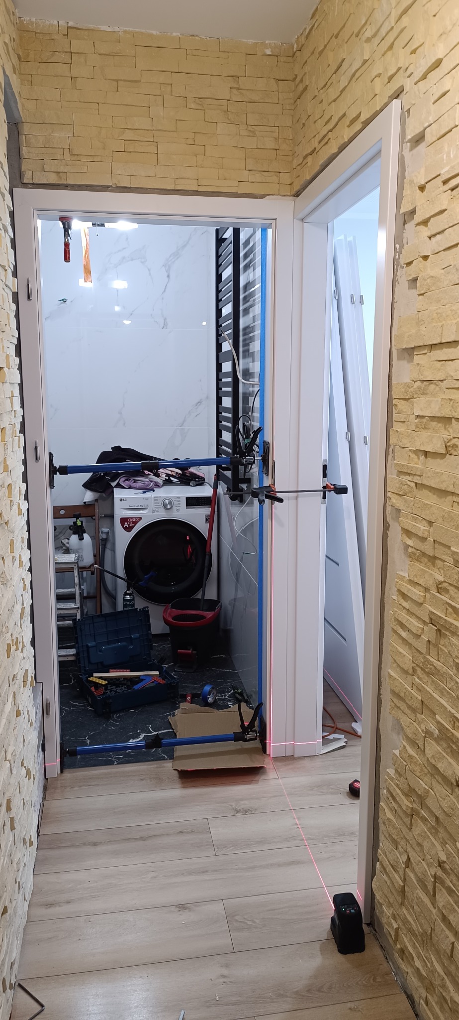 Montaż ościeżnicy drzwiowej w łazience z użyciem poziomicy laserowej. Widoczne narzędzia i przymocowana ościeżnica. Ściana wykończona kamieniem dekoracyjnym.