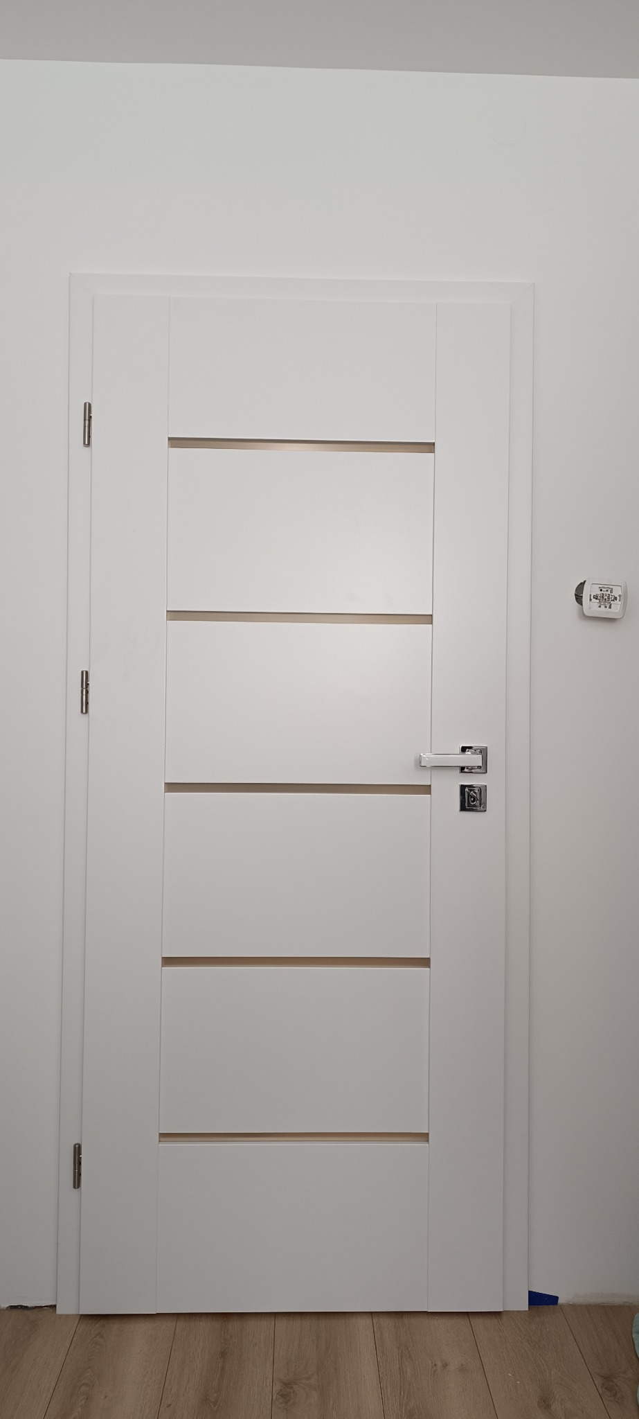 Białe drzwi wewnętrzne z poziomymi frezami i chromowaną klamką, zamontowane w białej ścianie. Widoczny fragment podłogi z paneli.