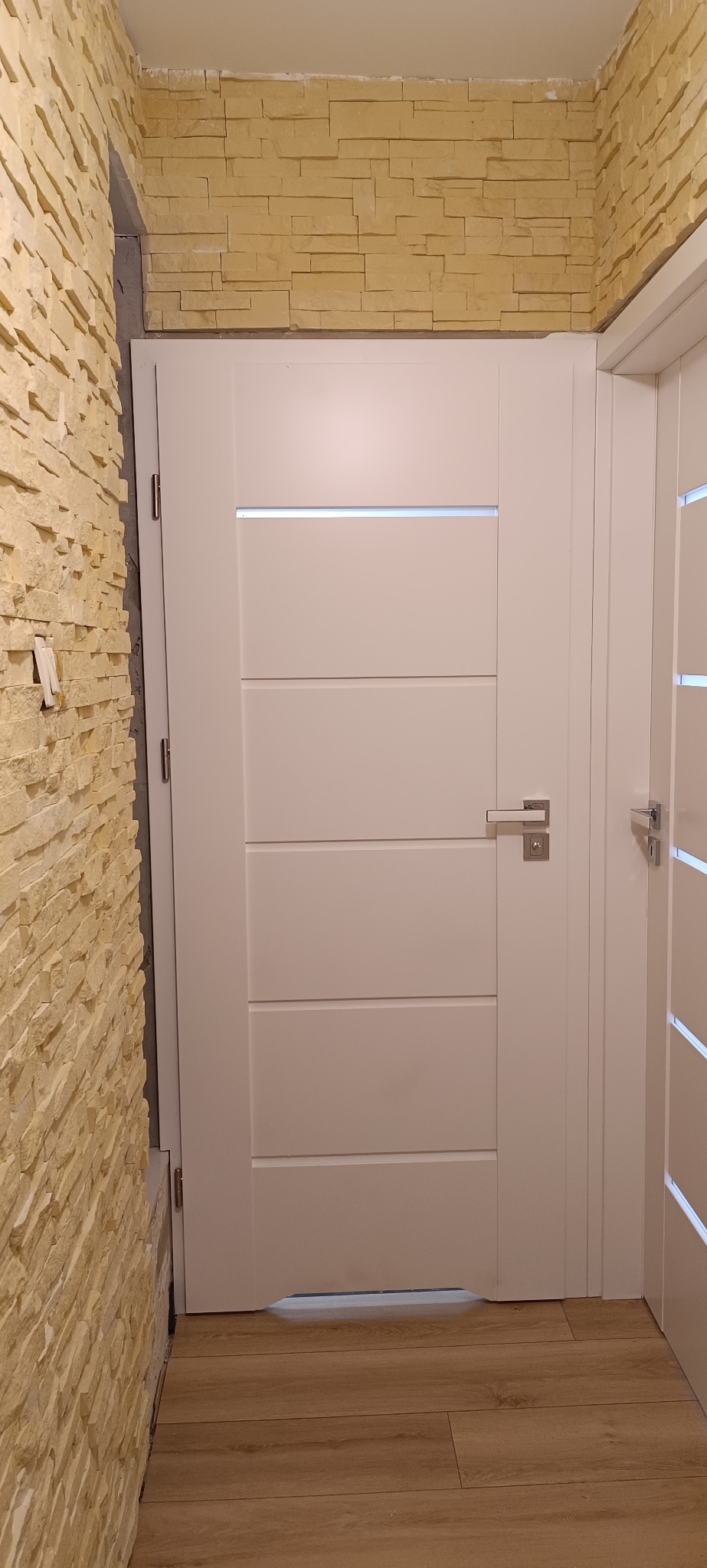 Białe drzwi wewnętrzne z poziomymi panelami i przeszkleniem, zamontowane w ścianie wykończonej dekoracyjną cegłą. Widoczny fragment sąsiednich drzwi.