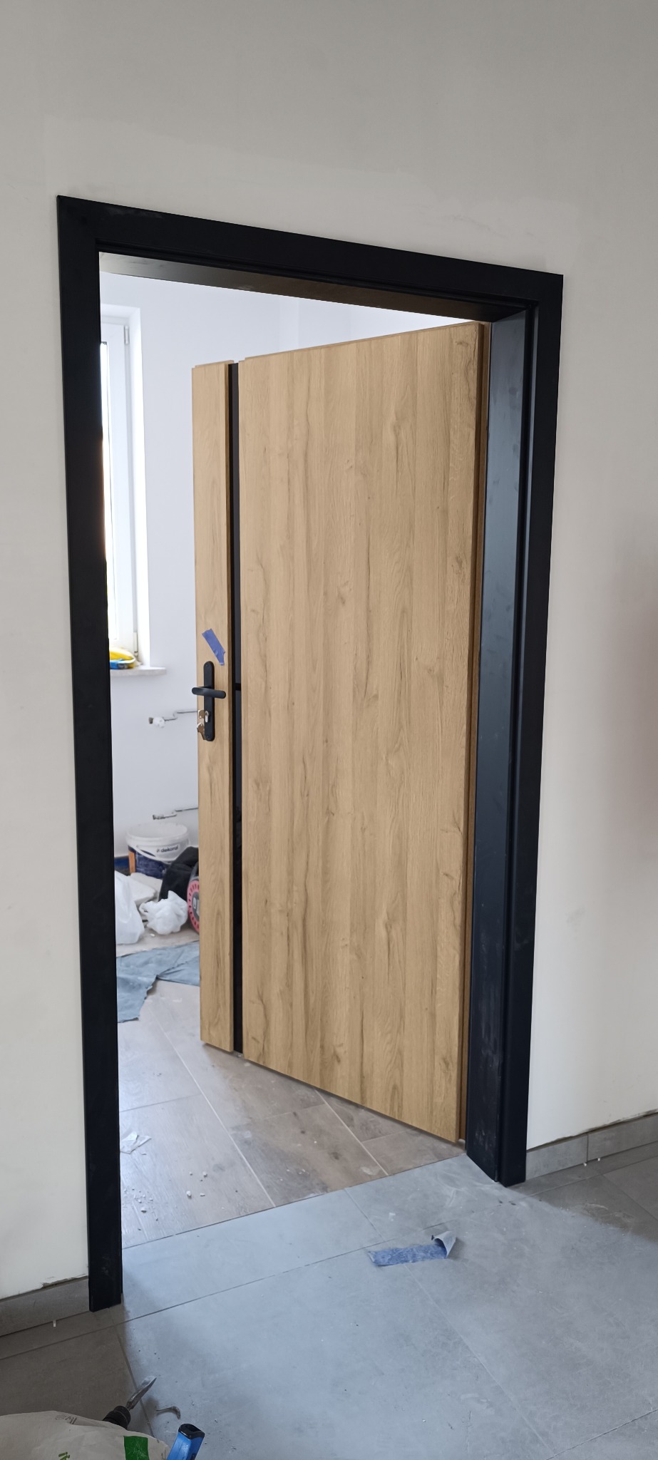 Częściowo otwarte drzwi wewnętrzne z drewnianą okleiną i czarną ościeżnicą. Widoczne fragmenty podłogi i ściany. W tle okno i przedmioty sugerujące prace wykończeniowe.