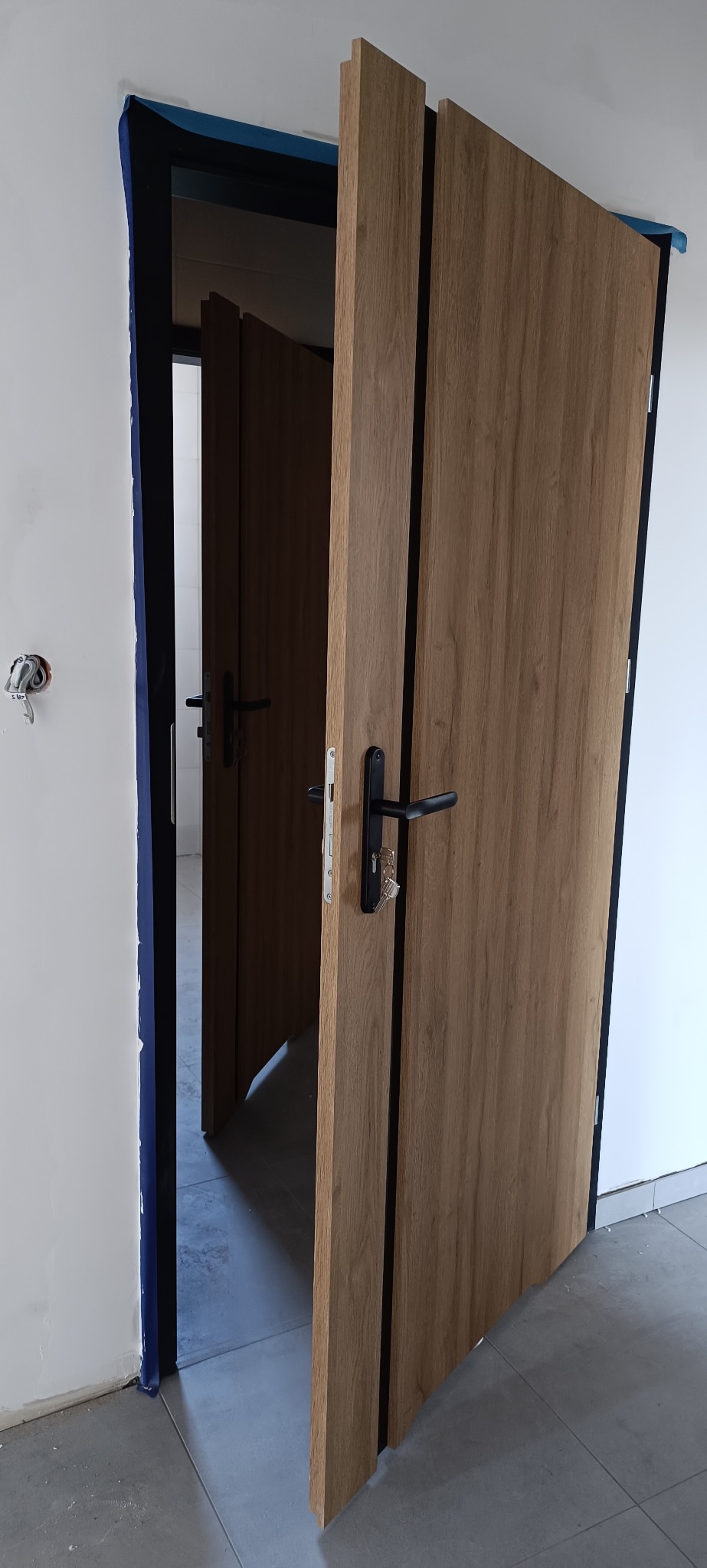 Otwarta, drewniana, nowoczesna drzwi wewnętrzne z czarną klamką, widoczne zabezpieczenia na ścianie, w tle kolejne drzwi. Minimalistyczny design.