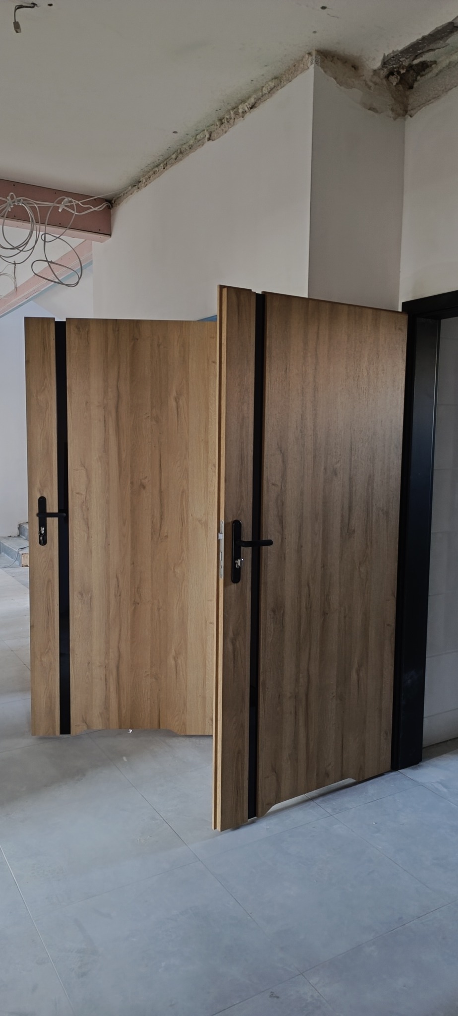 Dwie drewnopodobne drzwi wewnętrzne z czarnymi akcentami, jedna otwarta, druga zamknięta, na tle surowego wnętrza w trakcie remontu.