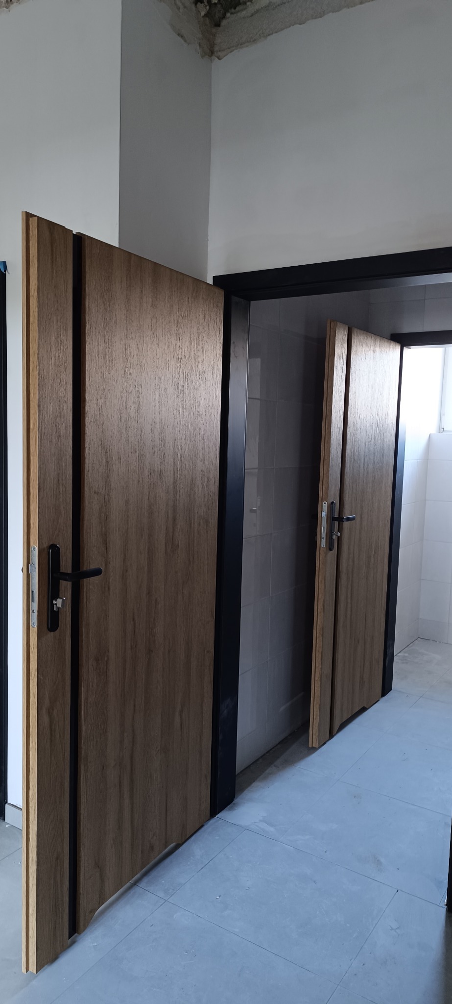 Dwie drewnopodobne drzwi wewnętrzne z czarnymi ościeżnicami, jedna otwarta, ukazująca wykończoną łazienkę z białymi płytkami. Widoczny fragment surowego sufitu.