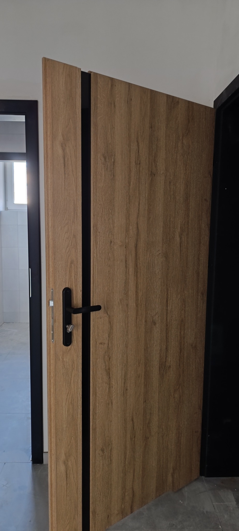 Otwarta, drewnopodobna drzwi wewnętrzne z czarną klamką i ościeżnicą, widok na jasne pomieszczenie z białymi płytkami. Minimalistyczny design.