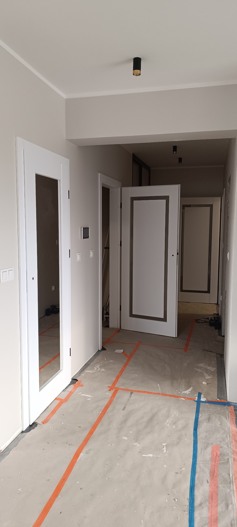 Wnętrze w trakcie remontu z widocznymi białymi drzwiami z przeszkleniami i bez, zabezpieczona podłoga folią malarską. Widok na kilka pomieszczeń.