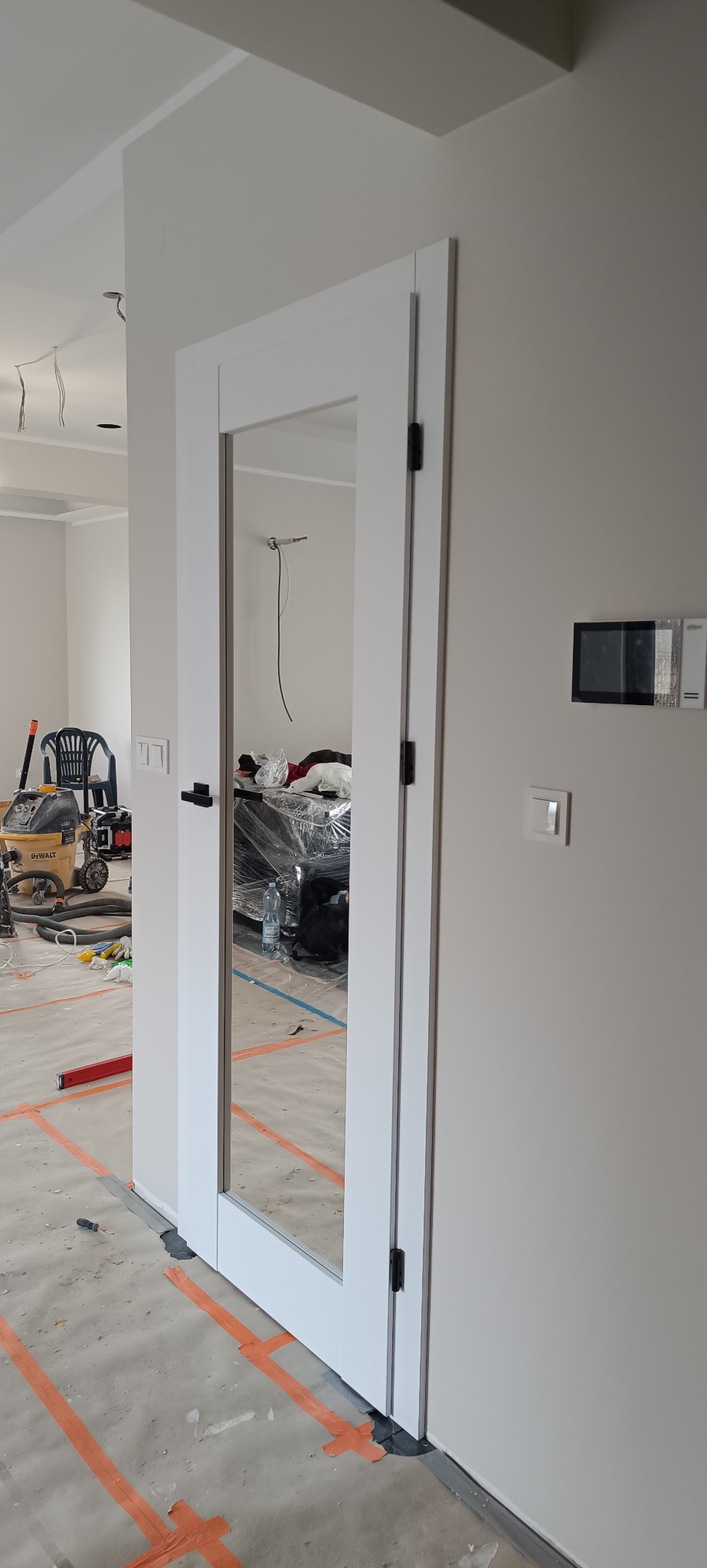 Białe, nowoczesne drzwi wewnętrzne z szybą, zamontowane w surowym wnętrzu. Widoczne narzędzia i zabezpieczenia podłogi, co sugeruje etap wykończeniowy.