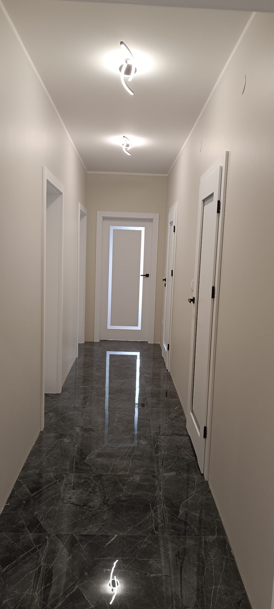 Korytarz z szarą marmurową podłogą i białymi drzwiami z czarnymi klamkami. Nowoczesne oświetlenie sufitowe. Perspektywa z głębi korytarza.