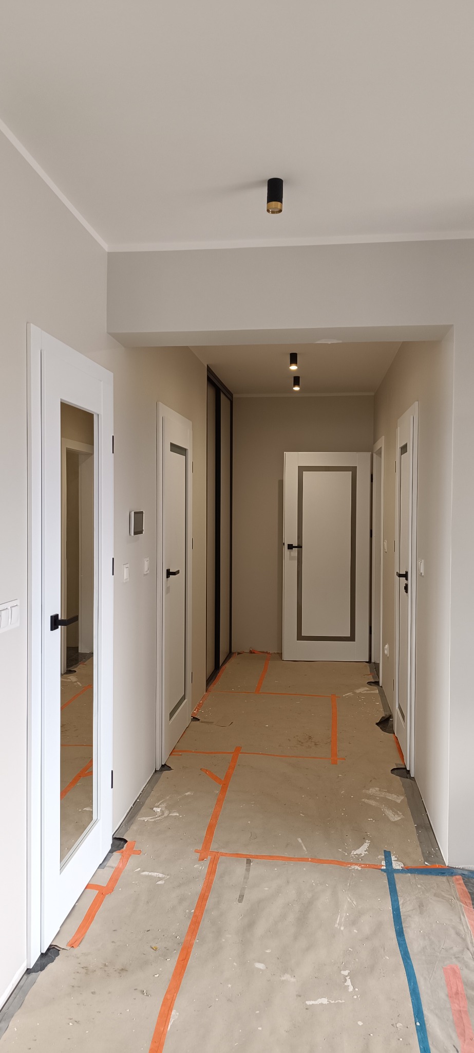 Korytarz w trakcie remontu z widocznymi białymi drzwiami z czarnymi klamkami i zabezpieczoną podłogą. Widoczne lampy sufitowe.