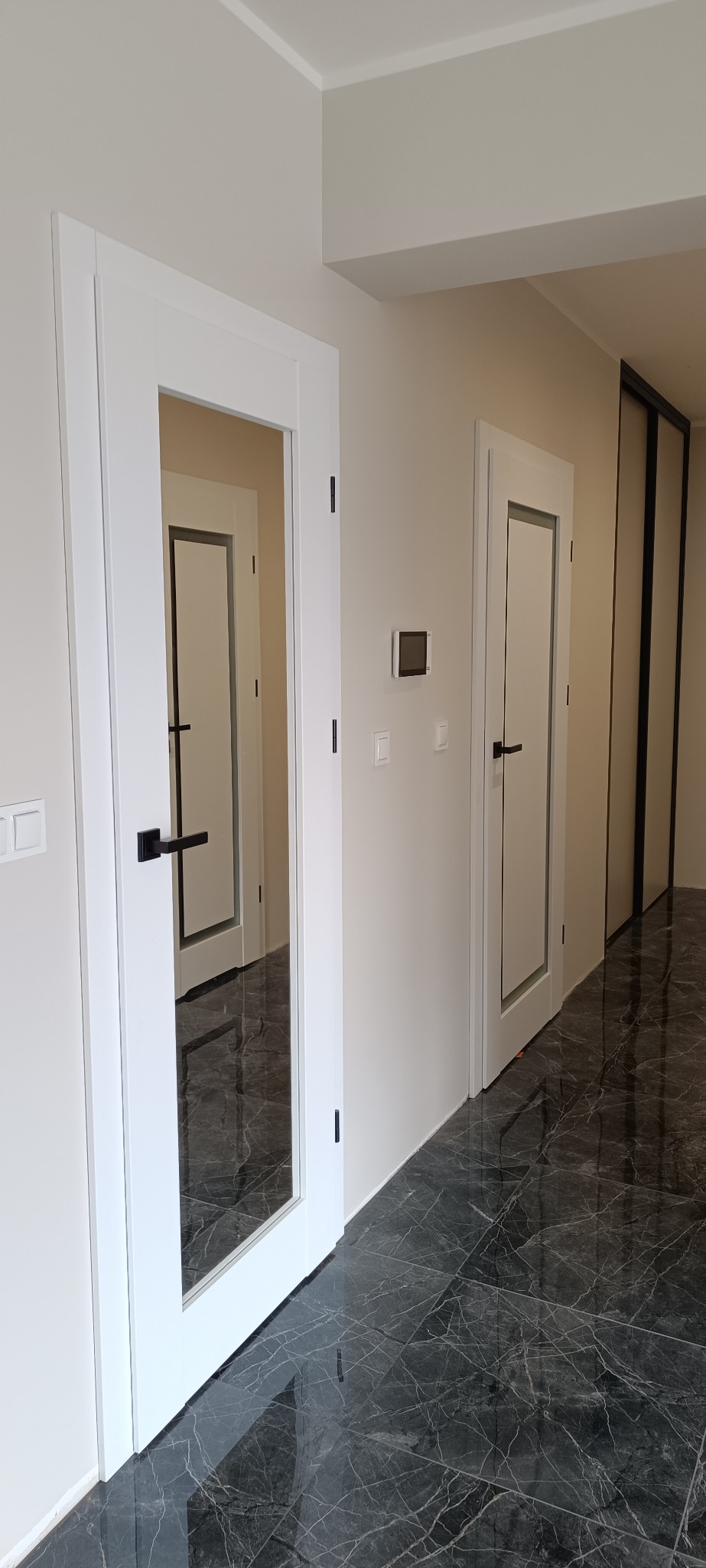 Białe, przeszklone drzwi wewnętrzne z czarną klamką, widoczne w korytarzu z marmurową podłogą. W tle kolejne drzwi i przesuwne drzwi szafy.