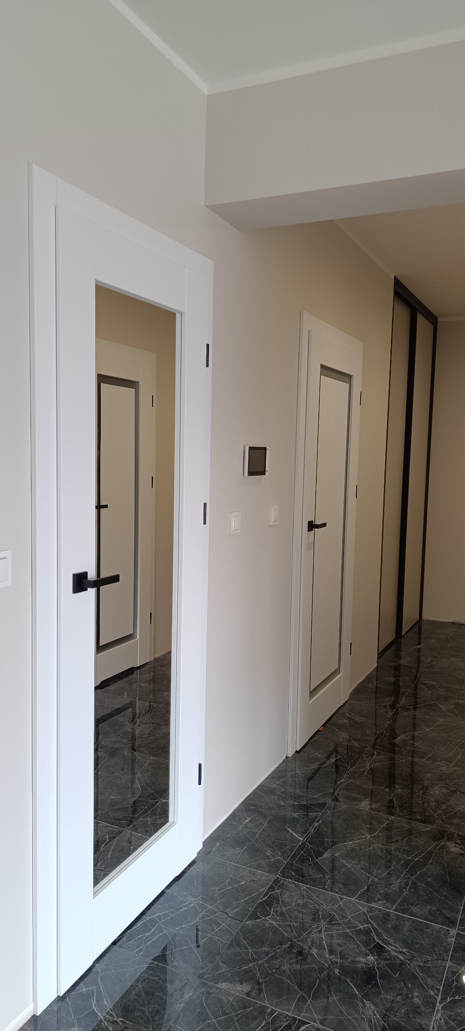 Nowoczesne, białe drzwi wewnętrzne z czarnymi klamkami, widoczne w perspektywie korytarza z marmurową podłogą. Minimalistyczny design, jasne ściany.
