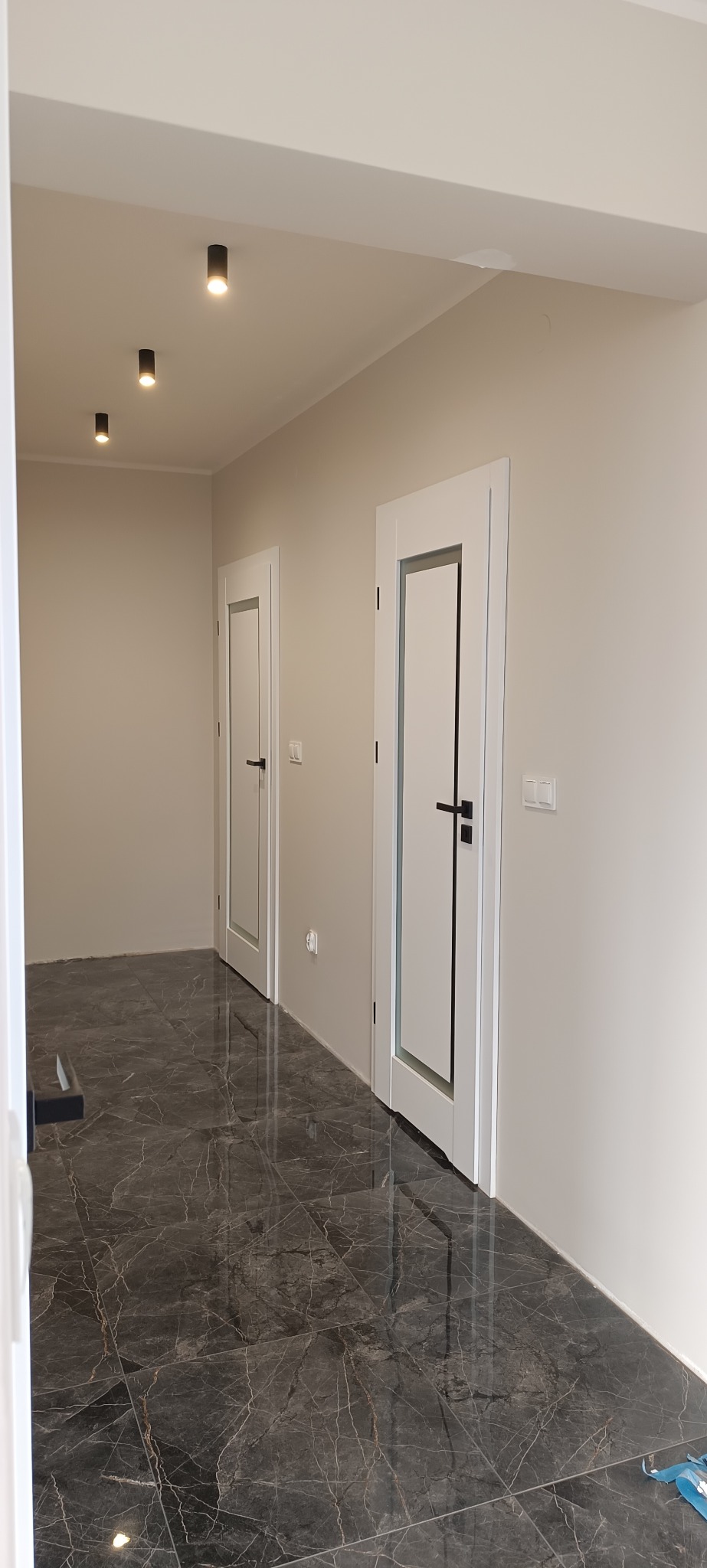 Nowoczesne, białe drzwi wewnętrzne z czarnymi klamkami w korytarzu z ciemną, marmurową podłogą. Minimalistyczny design, punktowe oświetlenie sufitowe.
