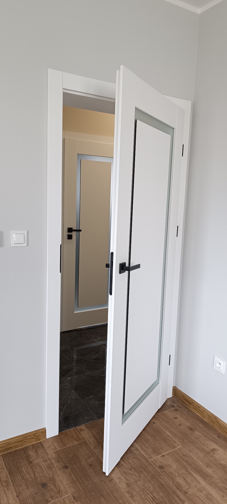 Białe, częściowo otwarte drzwi wewnętrzne z czarną klamką, widok na kolejne drzwi i marmurową podłogę w głębi. Jasne ściany, drewnopodobna podłoga.