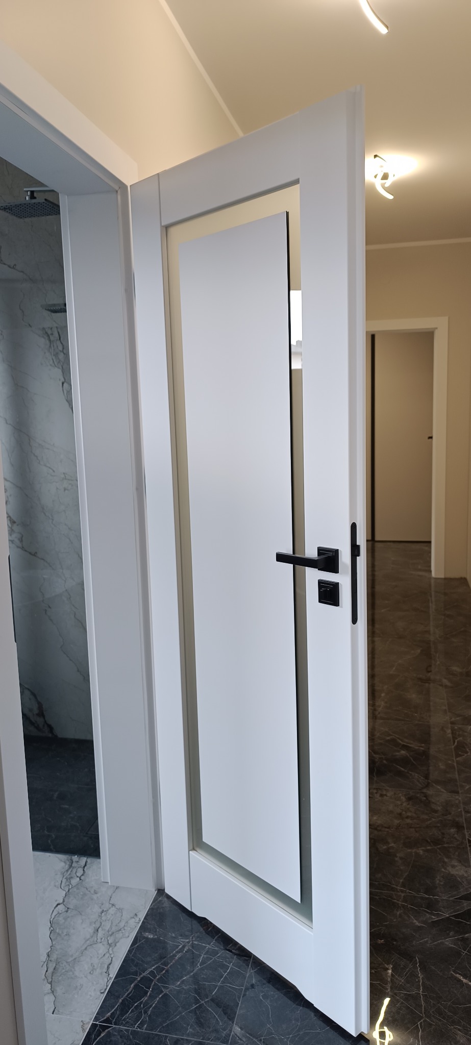 Nowoczesne, białe drzwi wewnętrzne z czarną klamką, częściowo otwarte, ukazujące łazienkę z marmurowymi płytkami i dalsze pomieszczenia. Minimalistyczny design, jasne wnętrze.