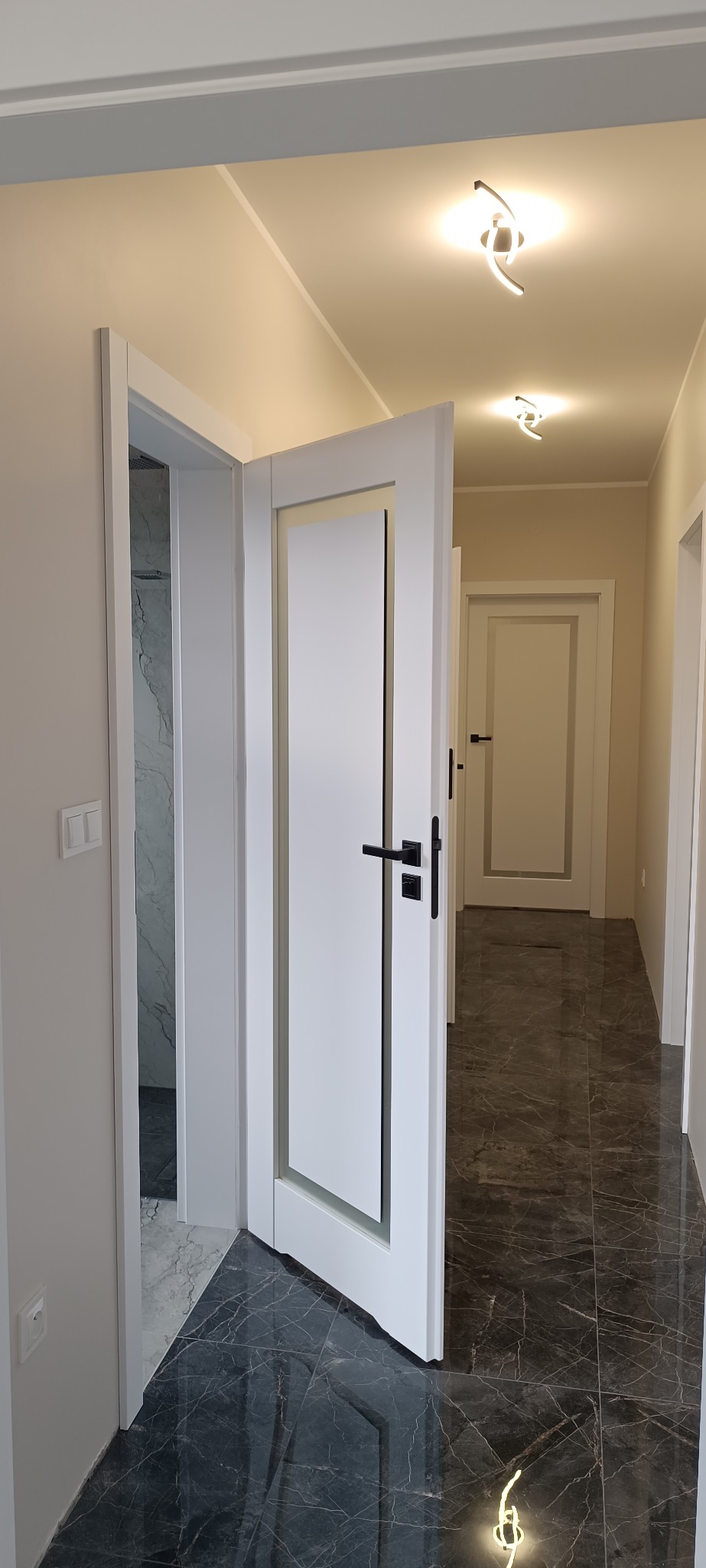 Białe, nowoczesne drzwi wewnętrzne z czarną klamką, otwarte w jasnym korytarzu z marmurową podłogą. Widoczne kolejne drzwi w głębi. Minimalistyczne oświetlenie sufitowe.