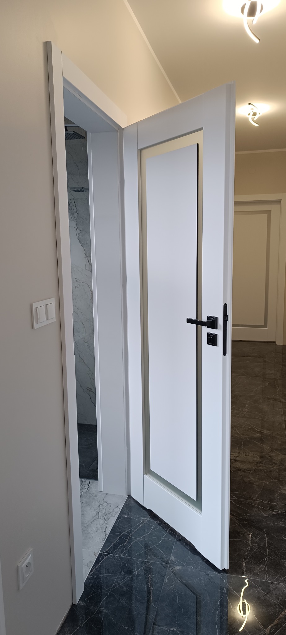 Nowoczesne, białe drzwi wewnętrzne z czarną klamką, otwarte do łazienki z marmurowymi płytkami. Widoczny fragment podłogi z ciemnego marmuru.