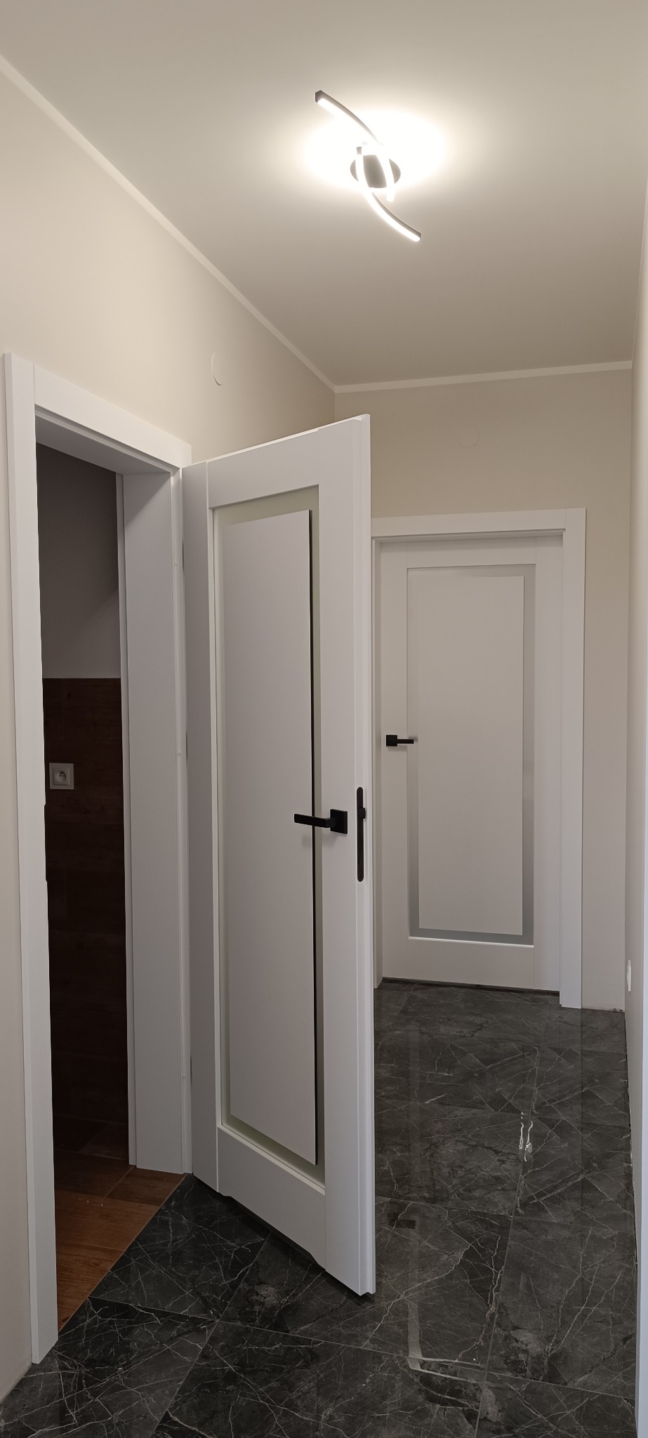 Nowoczesne, białe drzwi wewnętrzne z czarną klamką, częściowo otwarte, ukazujące wnętrze. W tle widoczne kolejne drzwi i marmurowa podłoga.