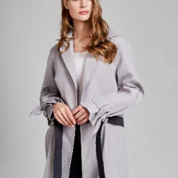 Kobieta z długimi, kręconymi włosami w szarym płaszczu z czarnym paskiem, pozuje na jasnoszarym tle.