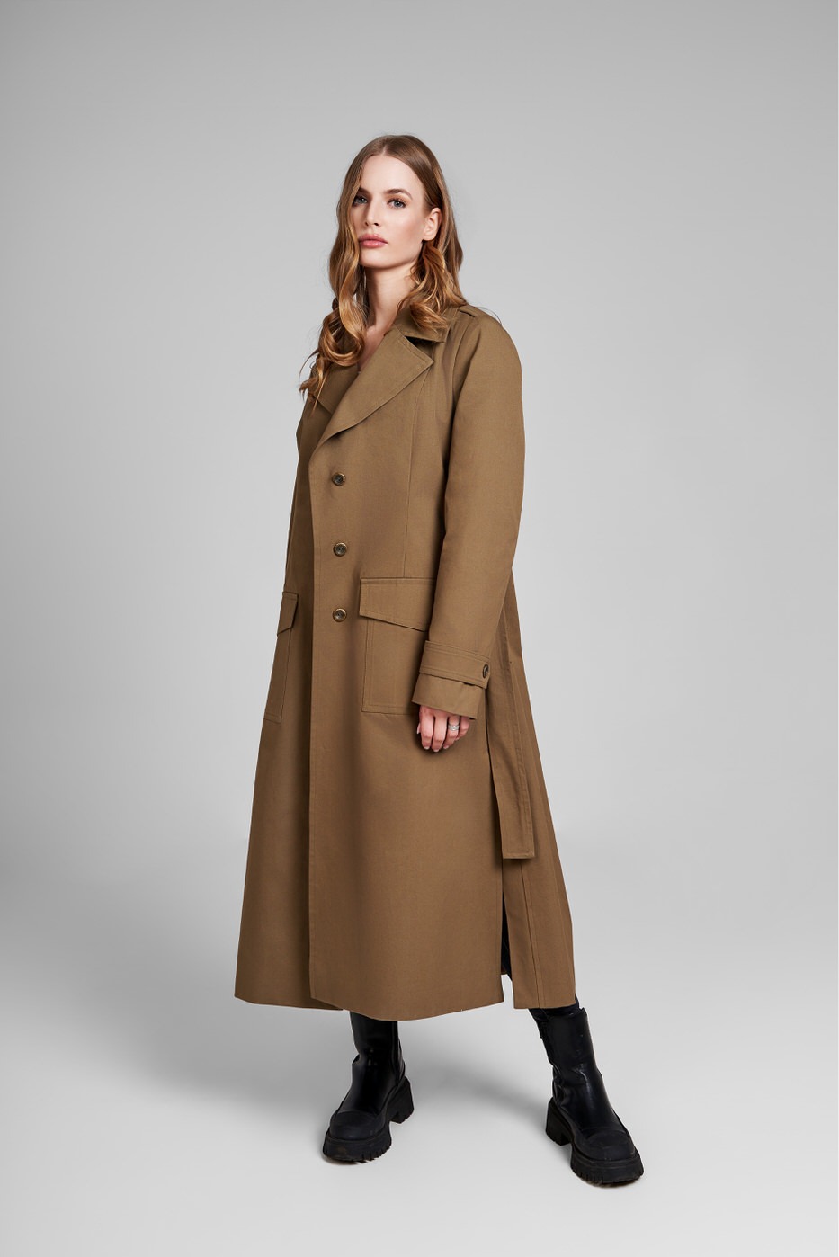 Kobieta w długim, brązowym płaszczu z szerokimi klapami i czarnych botkach na platformie, pozująca na jasnoszarym tle.
