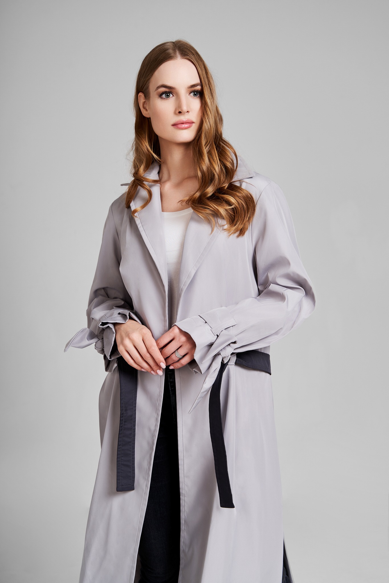 Kobieta z długimi, kręconymi włosami w szarym płaszczu z czarnym paskiem, pozuje na jasnoszarym tle.