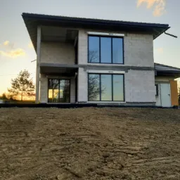 Surowy stan dwupiętrowego domu z betonu komórkowego w trakcie budowy, widoczne okna z roletami zewnętrznymi, świeżo przekopana ziemia na pierwszym planie, zachodzące słońce w tle.