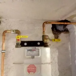 Zamontowany gazomierz z miedzianymi rurami i żółtymi zaworami odcinającymi na tle surowej, pomazanej ściany z widocznymi zaciekami i rurą wentylacyjną w izolacji.