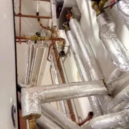 Złożona instalacja rur miedzianych i izolowanych, widoczne zawory, manometry oraz fragment pieca, całość zamontowana na białej ścianie.