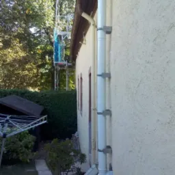 Biała rura spustowa zamontowana na elewacji domu o kremowej strukturze, widoczne metalowe uchwyty mocujące, w tle rusztowanie i zieleń ogrodu.